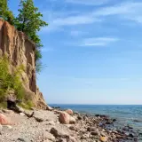 Klif Orłowski z zielenią na szczycie i kamienistą plażą u podnóża. Błękitne niebo i spokojne morze.