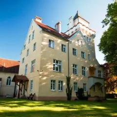 Zamek Nowęcin, żółty budynek z wieżą, otoczony zielenią i trawnikiem, w słoneczny dzień.