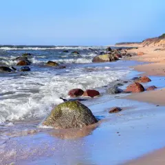 Poddąbie plaża. Fale rozbijają się o kamienie na piaszczystym brzegu. W oddali widać spacerującą parę.