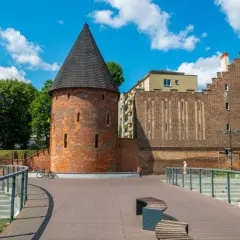 Kamienna baszta czarownic z stożkowym dachem, otoczona nowoczesną architekturą i zielenią, pod błękitnym niebem z chmurami.