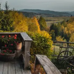 Drewniana zagroda Magoda z czerwonymi pelargoniami na werandzie. W tle jesienny krajobraz z pagórkami i kolorowymi drzewami.