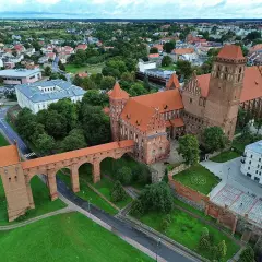 Zamek w Kwidzynie z charakterystycznym gdaniskiem i długim mostem nad fosą, otoczony zielenią i zabudowaniami miasta.