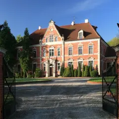 Pałac Leźno, okazała ceglana budowla z czerwonej cegły, otoczona zielenią, zaprasza przez otwartą, kutą bramę.