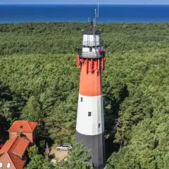 Wysoka, wielobarwna łeba latarnia morska góruje nad gęstym lasem sosnowym, z błękitnym morzem w tle.