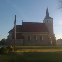 Stary kościół z cegły z wieżą i krzyżem przed fasadą, otoczony zielenią.
