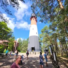 Wysoki, biało-czerwony **sasino stilo** otoczony sosnowym lasem. Grupa turystów zwiedza teren.