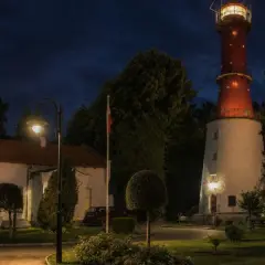 Wieczorem na przylądku na pobrzeżu kaszubskim widać oświetloną latarnię morską, budynki i ceglany komin.