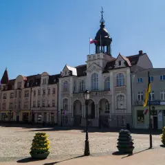 Słoneczny wejherowo rynek z ratuszem i kolorowymi budynkami. Na bruku stoją ozdobne donice z kwiatami.