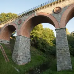 Imponujący, ceglany bytów most kolejowy z kamiennymi filarami i ozdobnymi medalionami, otoczony zielenią.