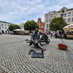 Pomnik pisarza na brukowanym kościerzyna rynek. W tle budynki, drzewa i ogródki kawiarniane.