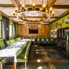 Elegancka sala bankietowa z długim stołem, zielonymi aksamitnymi krzesłami i stylowym, okrągłym żyrandolem. W tle zabytkowa komoda, a na ścianach ozdobne lustra. Idealne miejsce na spotkanie w stylu nordowi mol.