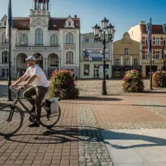 Mężczyzna na rowerze przejeżdża przez rynek przed ratuszem w Wejherowie. Słoneczny dzień, brukowana nawierzchnia, ozdobne krzewy.