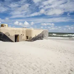 Betonowy bunkier z kamuflażem na piaszczystej plaży w Jastarni. Morze i niebo z chmurami w tle.