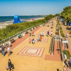 Tętniąca życiem ustka promenada w słoneczny dzień. Ludzie spacerują, odpoczywają na ławkach, a w tle widać plażę pełną turystów i morze.