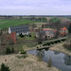 Zamek Kiszewski, otoczony polami i stawem, z wieżą i budynkami w trakcie renowacji.