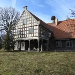 Stary dwór z muru pruskiego, z charakterystycznym gniazdem bociana na kominie. To historyczny lwi dwór, otoczony zielenią.