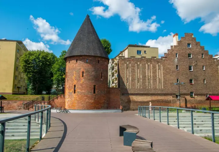 Kamienna baszta czarownic z stożkowym dachem, otoczona nowoczesną architekturą i zielenią, pod błękitnym niebem z chmurami.