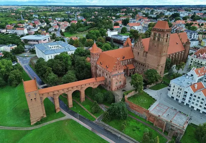 Zamek w Kwidzynie z charakterystycznym gdaniskiem i długim mostem nad fosą, otoczony zielenią i zabudowaniami miasta.