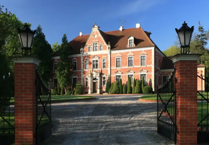 Pałac Leźno, okazała ceglana budowla z czerwonej cegły, otoczona zielenią, zaprasza przez otwartą, kutą bramę.