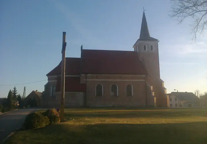 Stary kościół z cegły z wieżą i krzyżem przed fasadą, otoczony zielenią.