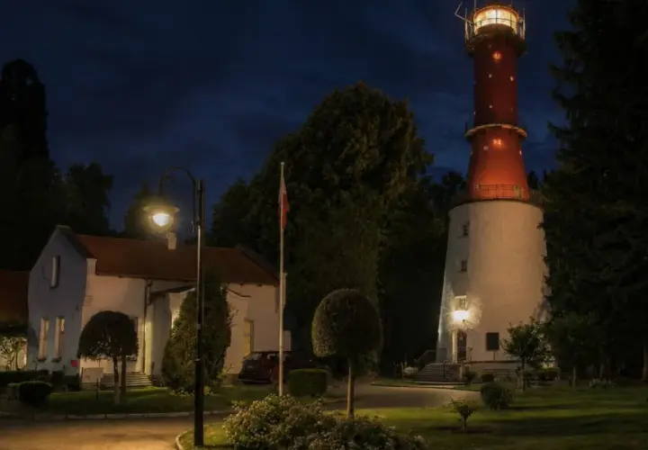 Wieczorem na przylądku na pobrzeżu kaszubskim widać oświetloną latarnię morską, budynki i ceglany komin.