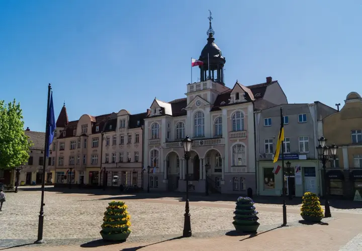 Słoneczny wejherowo rynek z ratuszem i kolorowymi budynkami. Na bruku stoją ozdobne donice z kwiatami.