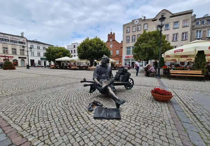 Pomnik pisarza na brukowanym kościerzyna rynek. W tle budynki, drzewa i ogródki kawiarniane.