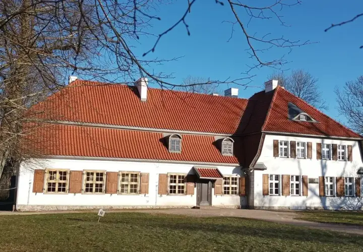 Dworek Wybickich z czerwonym dachem i białymi ścianami, otoczony nagimi drzewami, w słoneczny dzień.