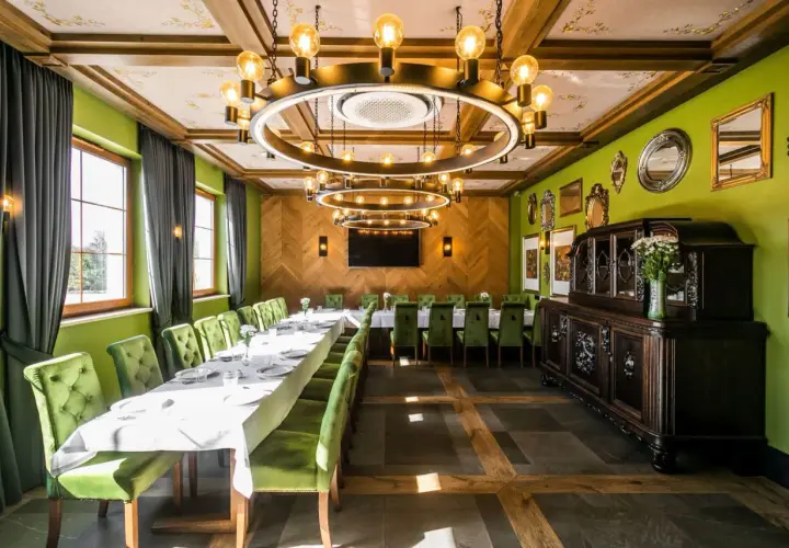 Elegancka sala bankietowa z długim stołem, zielonymi aksamitnymi krzesłami i stylowym, okrągłym żyrandolem. W tle zabytkowa komoda, a na ścianach ozdobne lustra. Idealne miejsce na spotkanie w stylu nordowi mol.