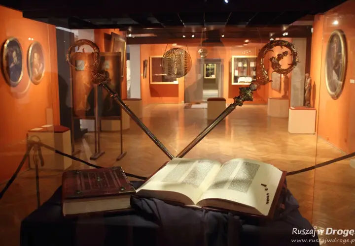 Otwarta biblia gutenberga pelplin, obok zamknięta księga i dwa ozdobne pastorały w muzeum.