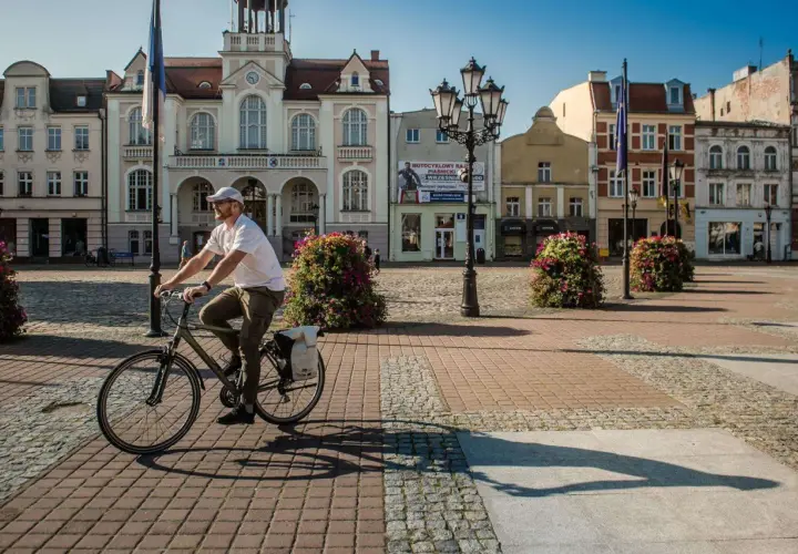 Mężczyzna na rowerze przejeżdża przez rynek przed ratuszem w Wejherowie. Słoneczny dzień, brukowana nawierzchnia, ozdobne krzewy.