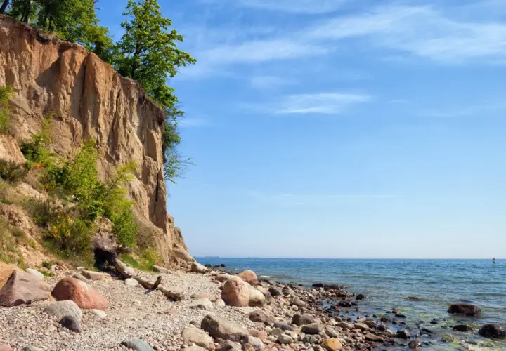 Klif Orłowski z zielenią na szczycie i kamienistą plażą u podnóża. Błękitne niebo i spokojne morze.