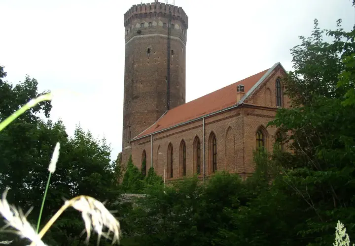 Zamek Człuchów, ceglana wieża i budynek z czerwonym dachem, otoczone zielenią drzew.
