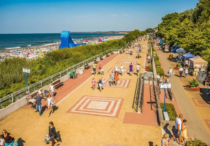 Tętniąca życiem ustka promenada w słoneczny dzień. Ludzie spacerują, odpoczywają na ławkach, a w tle widać plażę pełną turystów i morze.
