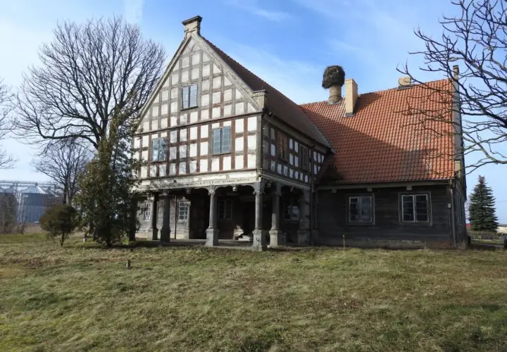Stary dwór z muru pruskiego, z charakterystycznym gniazdem bociana na kominie. To historyczny lwi dwór, otoczony zielenią.