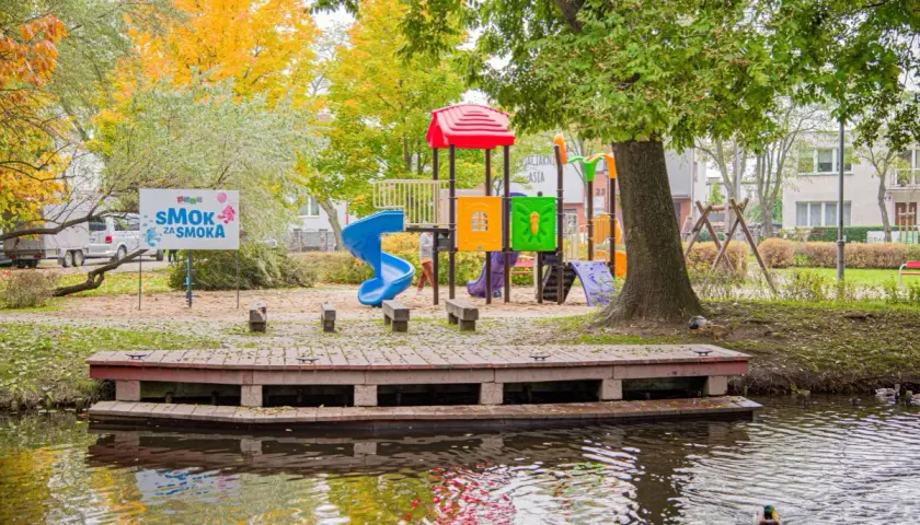Kolorowy plac zabaw i pomost nad wodą w Rumi. Jesienne liście i tablica "SMOK ZA SMOKA" zapraszają do zabawy.
