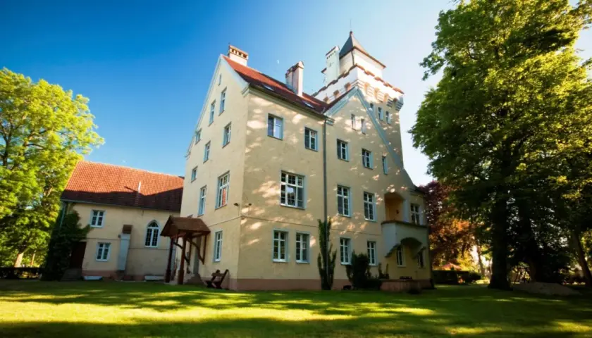 Zamek Nowęcin, żółty budynek z wieżą, otoczony zielenią i trawnikiem, w słoneczny dzień.