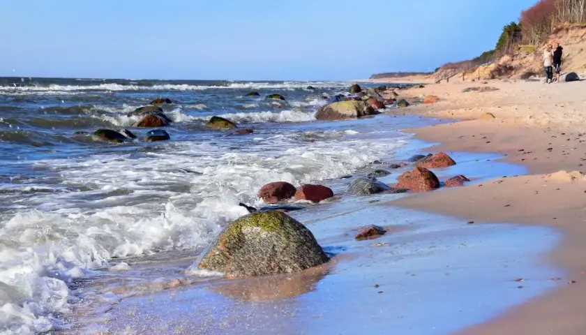 Poddąbie plaża. Fale rozbijają się o kamienie na piaszczystym brzegu. W oddali widać spacerującą parę.