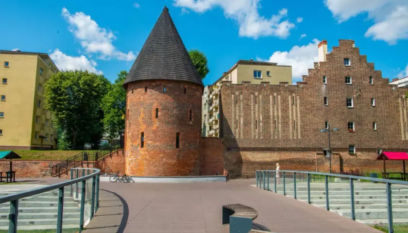 Kamienna baszta czarownic z stożkowym dachem, otoczona nowoczesną architekturą i zielenią, pod błękitnym niebem z chmurami.