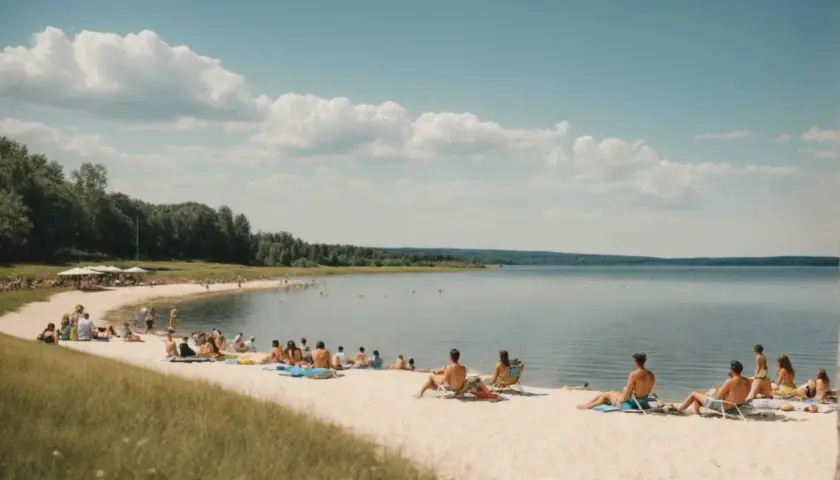 Ludzie odpoczywają na piaszczystej plaży w **Ostrzyce Park**. Woda jest spokojna, a niebo błękitne z białymi chmurami.
