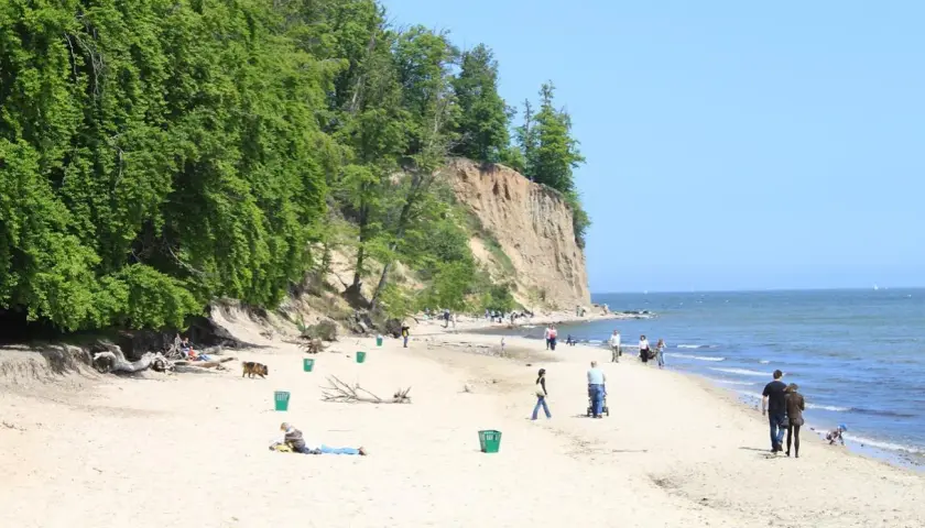 Piękny dzień na kąpieliskach Gdynia. Ludzie spacerują po piaszczystej plaży u podnóża zielonego klifu, ciesząc się słońcem i morzem.