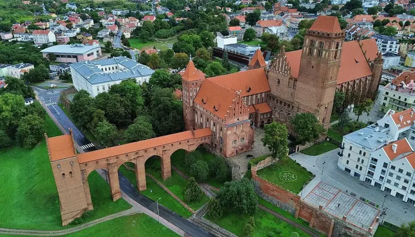 Zamek w Kwidzynie z charakterystycznym gdaniskiem i długim mostem nad fosą, otoczony zielenią i zabudowaniami miasta.