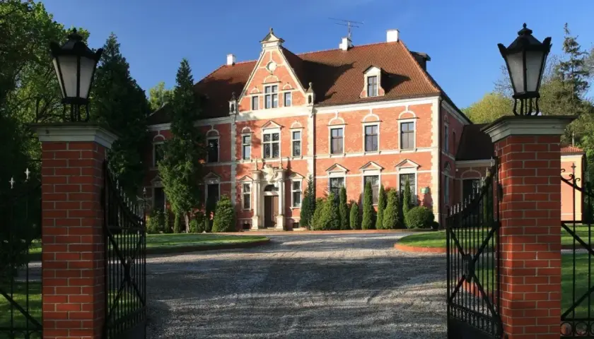 Pałac Leźno, okazała ceglana budowla z czerwonej cegły, otoczona zielenią, zaprasza przez otwartą, kutą bramę.