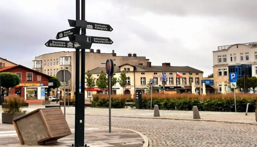 Kartuzy rynek z drogowskazami do kolegiaty, muzeum, dworca i informacji turystycznej. Na pierwszym planie przewrócony kosz.