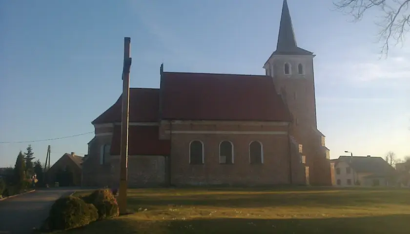 Stary kościół z cegły z wieżą i krzyżem przed fasadą, otoczony zielenią.