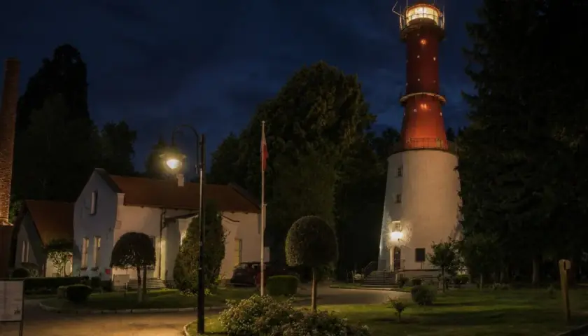 Wieczorem na przylądku na pobrzeżu kaszubskim widać oświetloną latarnię morską, budynki i ceglany komin.