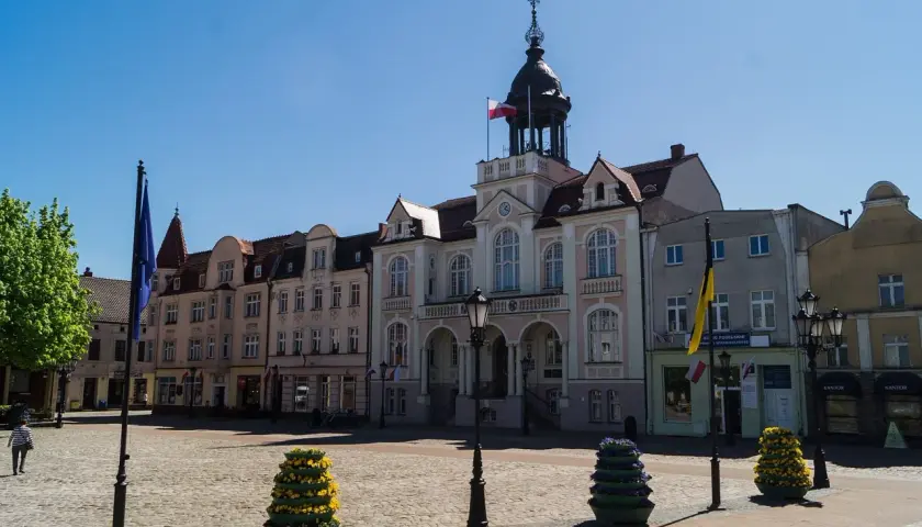 Słoneczny wejherowo rynek z ratuszem i kolorowymi budynkami. Na bruku stoją ozdobne donice z kwiatami.