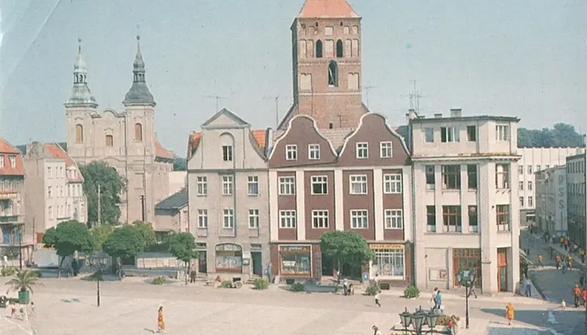 Chojnice rynek z kościołem i kamienicami. Ludzie spacerują po placu, tworząc tętniące życiem tło dla zabytkowej architektury.