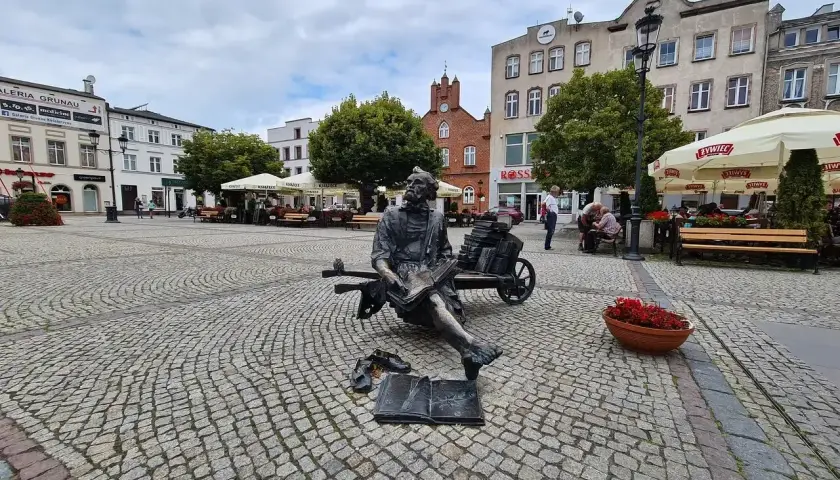 Pomnik pisarza na brukowanym kościerzyna rynek. W tle budynki, drzewa i ogródki kawiarniane.