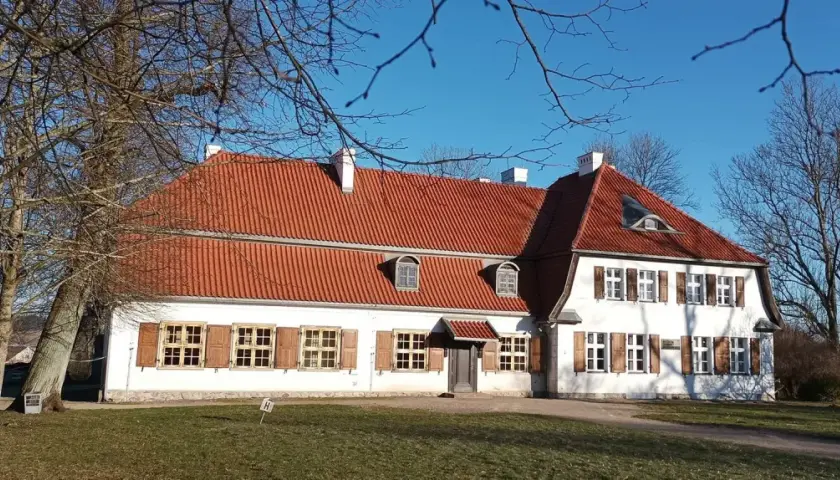 Dworek Wybickich z czerwonym dachem i białymi ścianami, otoczony nagimi drzewami, w słoneczny dzień.